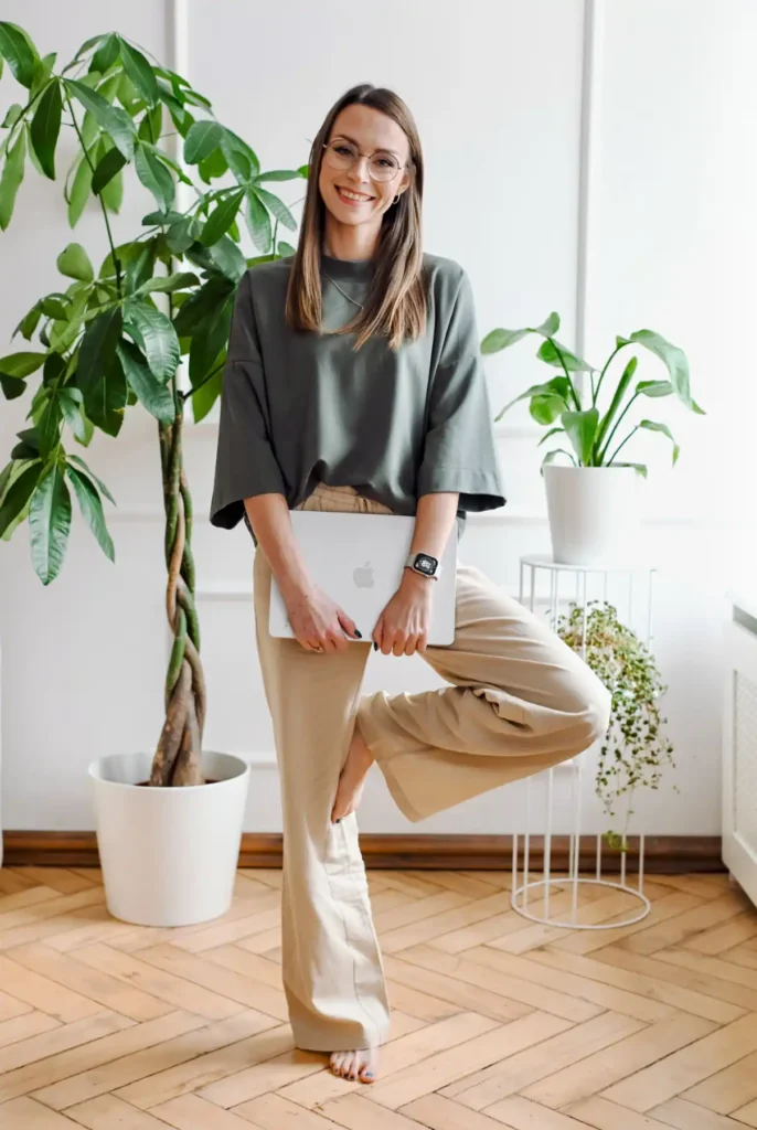 Psycholog stojąca z laptopem w jasnym wnętrzu z roślinami, uśmiechnięta i zrelaksowana.